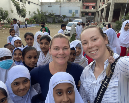 Two women smiling with a group of children outdoors in a courtyard