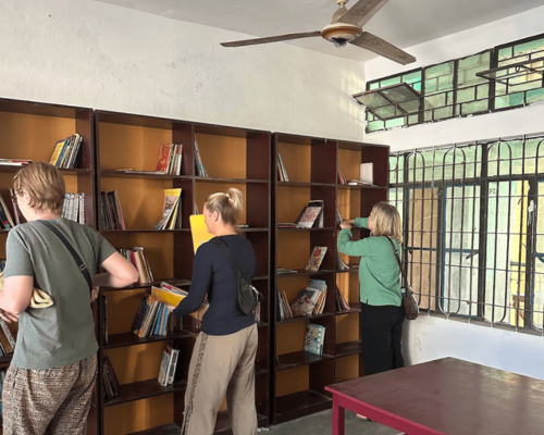 People browsing books in a wooden library room with shelves, windows, and a ceiling fan.