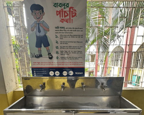 Public handwashing station with three faucets beneath a Bengali hygiene poster, in a school-like corridor.
