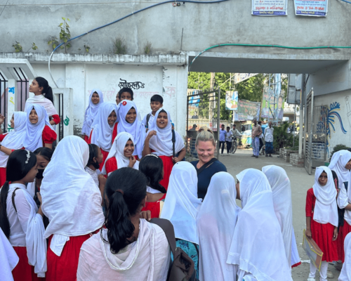 Students in white headscarves and red skirts gathered in a school courtyard near an archway.