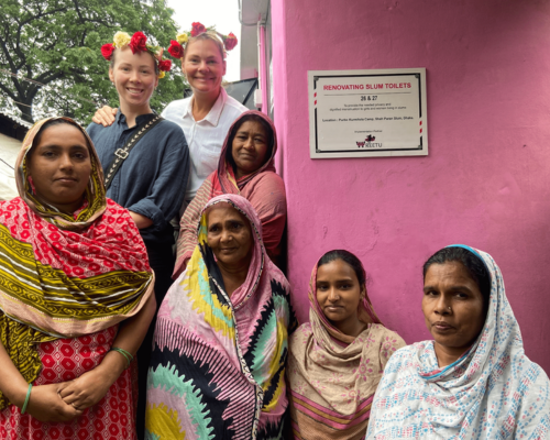 Seven women stand by a pink wall with a plaque; two wear flower crowns and smile.