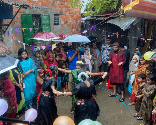 People dancing in a rainy courtyard during a lively neighborhood celebration, with umbrellas and balloons nearby.
