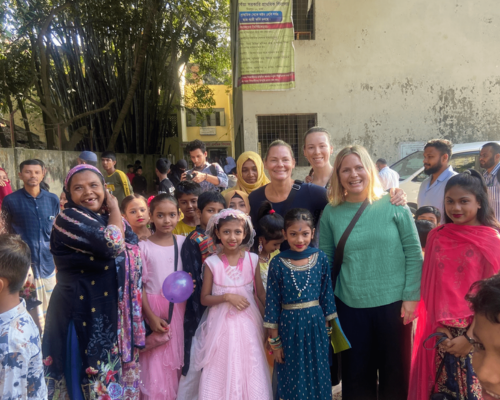 Smiling group posing outdoors, with children in colorful dresses and adults behind them.