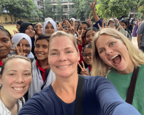 Smiling group selfie outdoors in a school courtyard, with students and three adults posing together