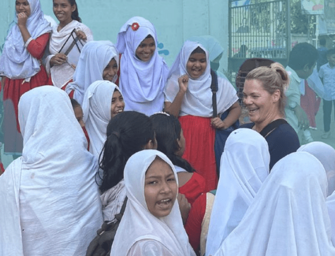 Smiling students in white headscarves gather around a woman outdoors in a school setting.