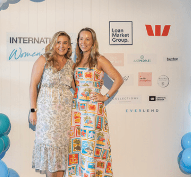 Two women smiling at a branded event backdrop, one in a white floral dress and one in a colorful patterned dress