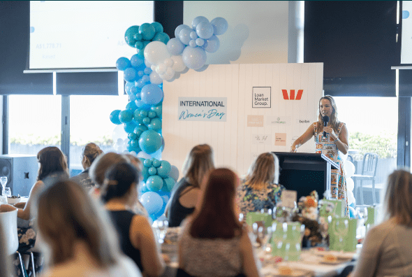 Speaker at podium addressing audience at a decorated event with blue-and-white balloon arch