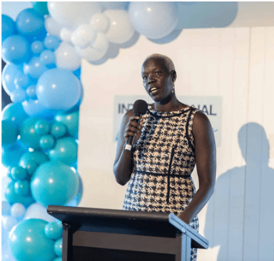 Speaker at podium holding microphone beside a blue and white balloon arch at an indoor event