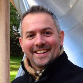 Smiling man in a black jacket outdoors, with a metallic sculpture in the background