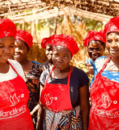 Group of smiling people in red aprons and headscarves standing outdoors under a thatched roof