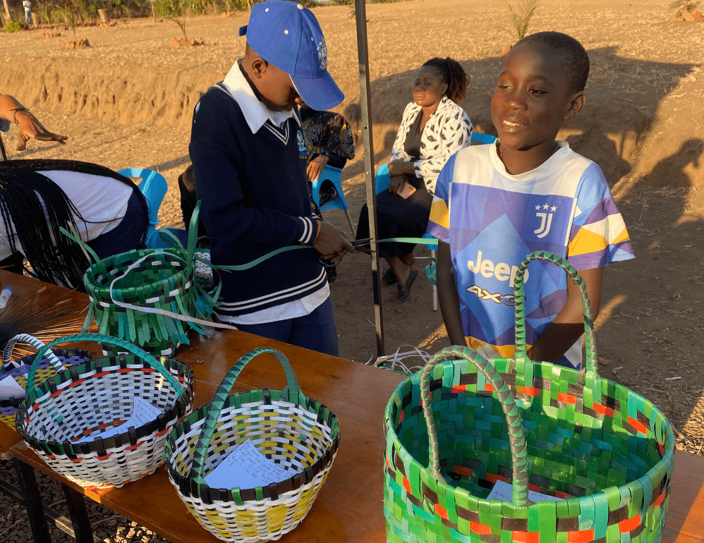 Children browsing colorful woven baskets at an outdoor market stall under bright sun.