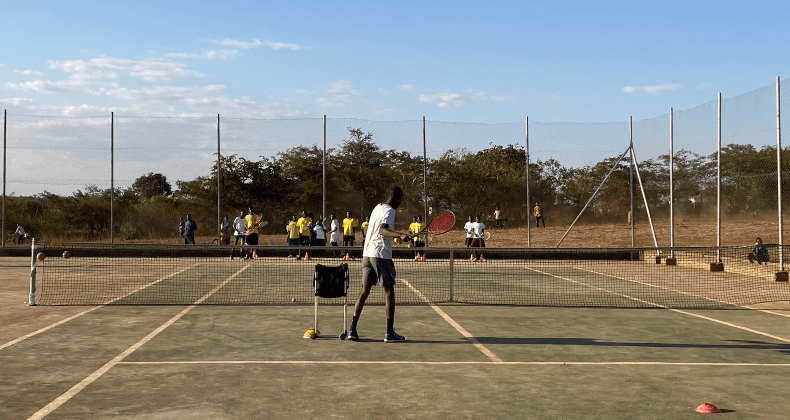 Tennis court with players practicing under a clear sky, one person in the foreground near the net.