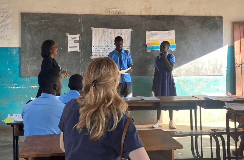 Students present at the front of a classroom while classmates watch from desks.