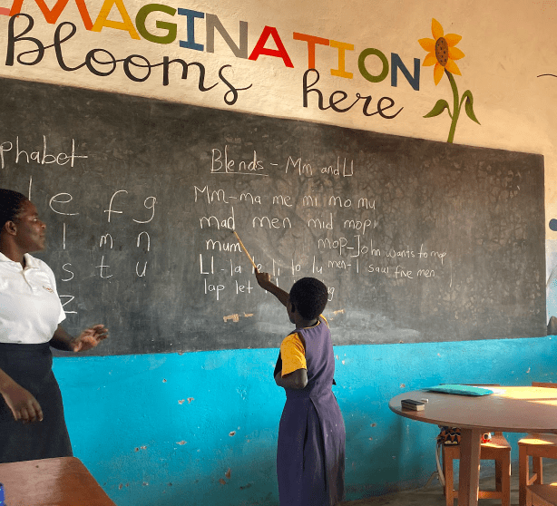 Teacher and student at a chalkboard in a classroom with “IMAGINATION Blooms here” painted above.