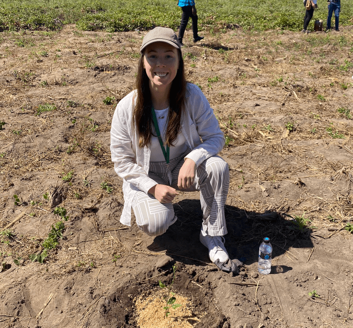Woman crouching in a dry field, smiling at the camera, with a water bottle nearby.