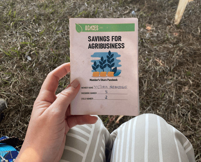 Hand holding a pink booklet titled “Savings for Agribusiness” while sitting on grass outdoors