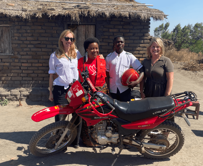 Four people standing beside a red dirt bike in front of a brick hut