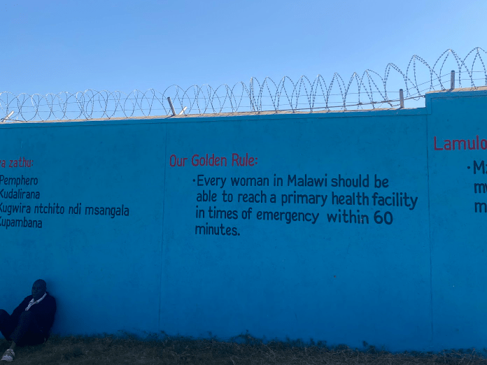 Blue wall with barbed wire and a health message; a person sits at the lower left.