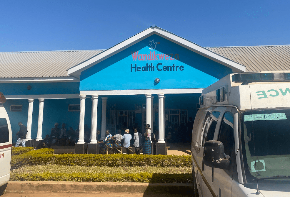Blue-roofed health centre entrance with people gathered outside and vehicles parked in front
