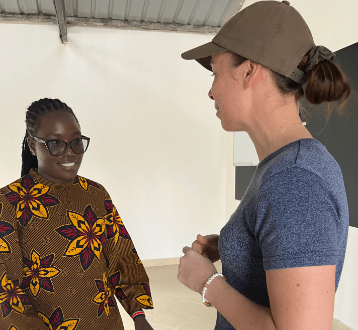 Two people talking indoors, one in a patterned dress and glasses, the other wearing a cap and blue shirt.