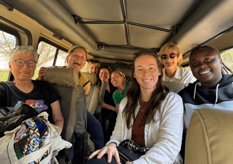 Six people smiling inside a vehicle, seated together with tan seats and a beige ceiling.