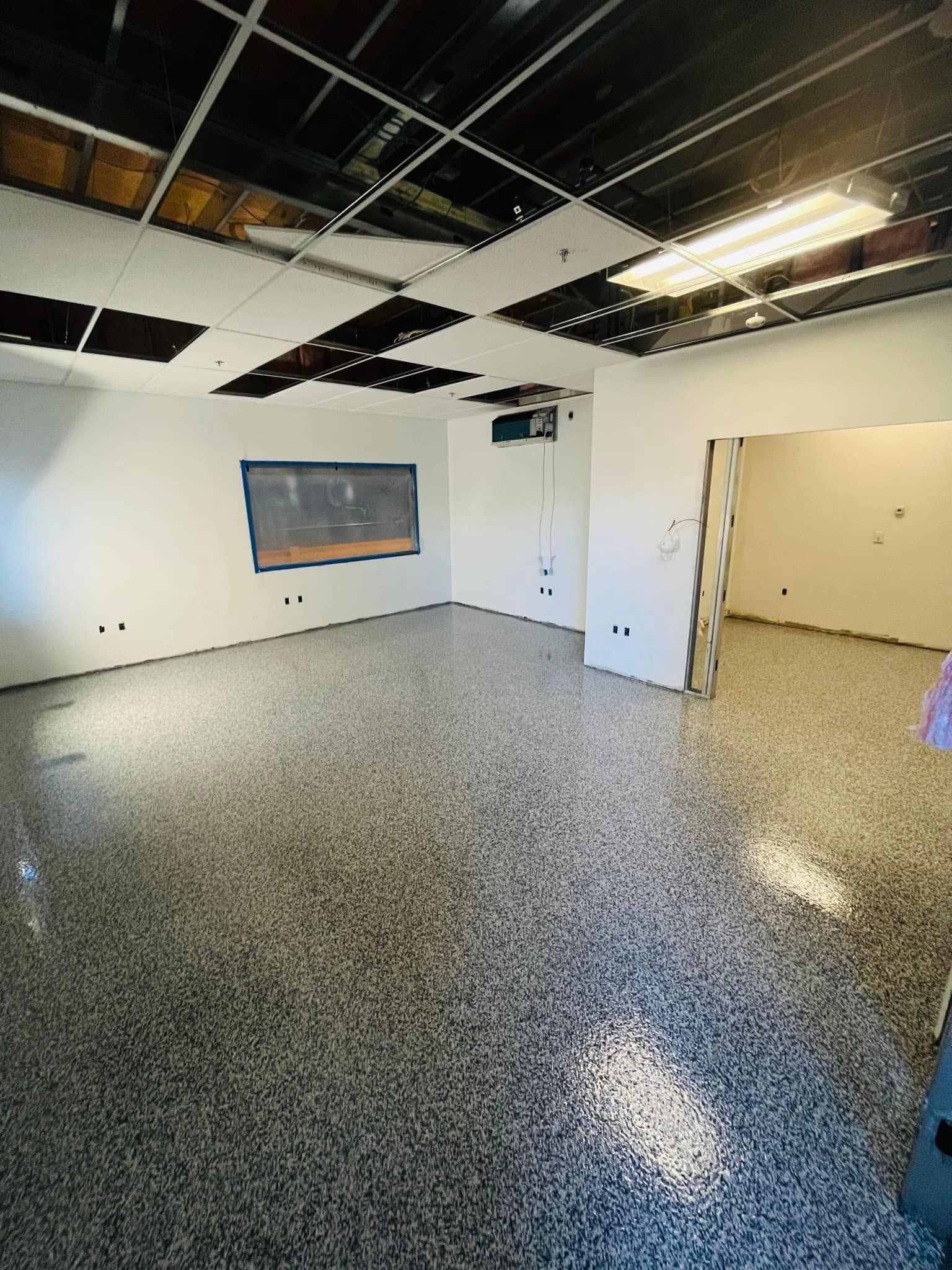 Empty office room with speckled floor, exposed ceiling tiles, white walls, and a window.