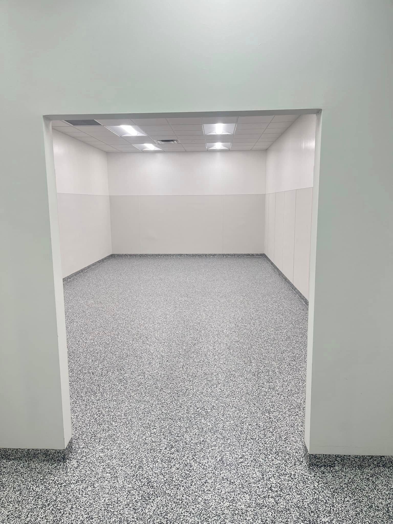 Empty white hallway leading to a small room with speckled gray carpet and ceiling lights