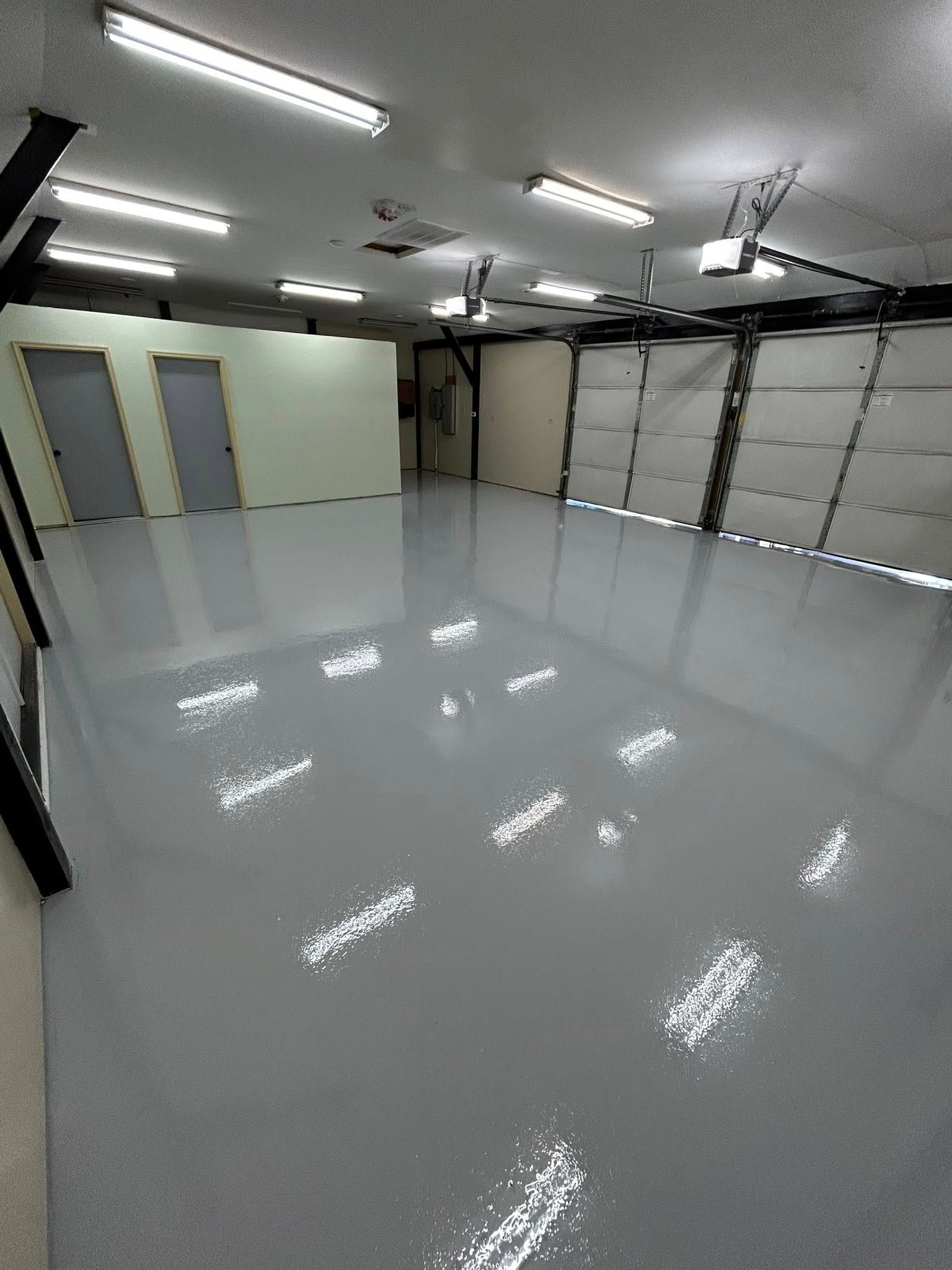 Empty polished garage with gray floor, white walls, overhead fluorescent lights, and closed garage doors.