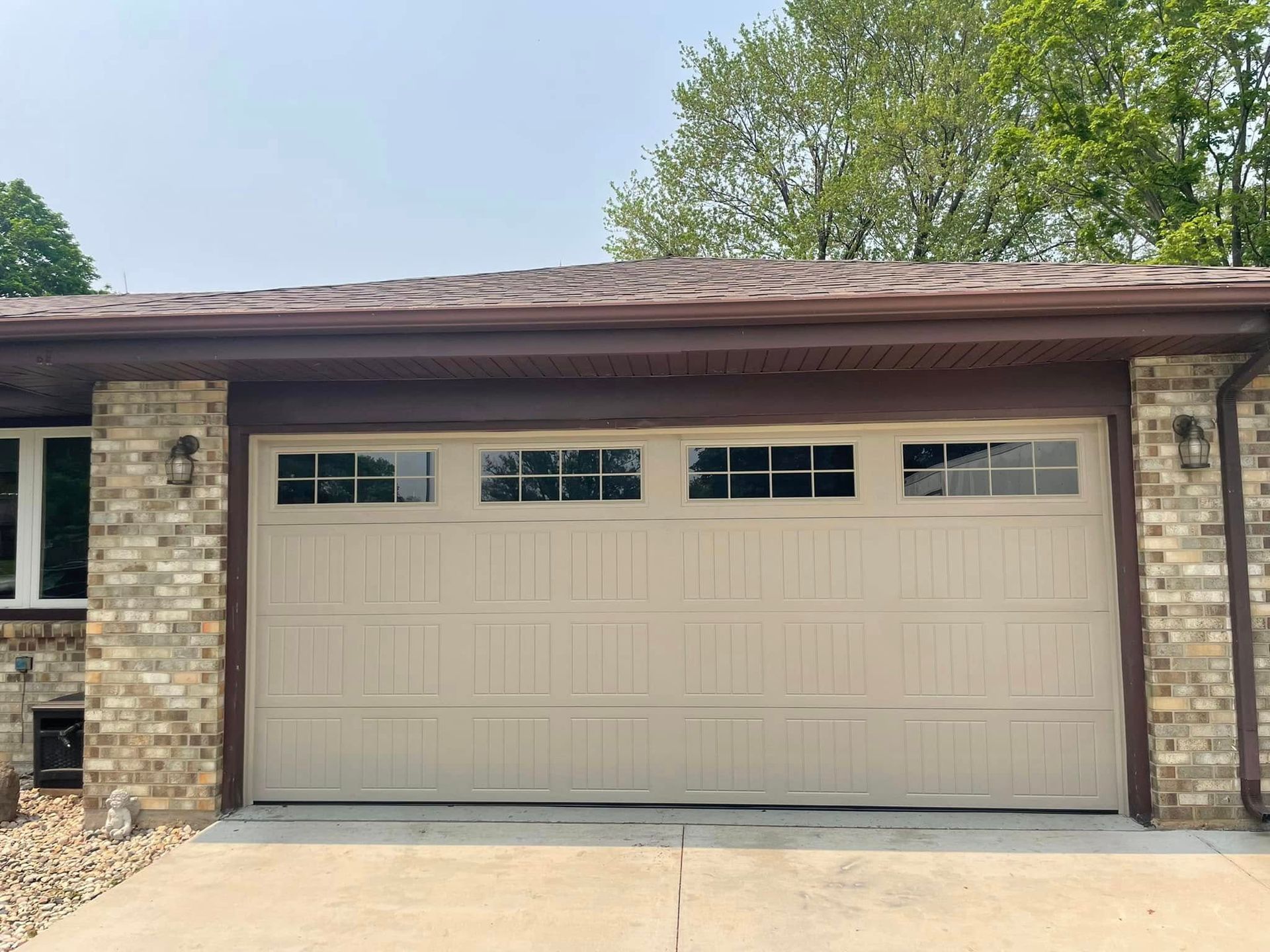 Beige two-car garage door on a brick house with small windows and trees above