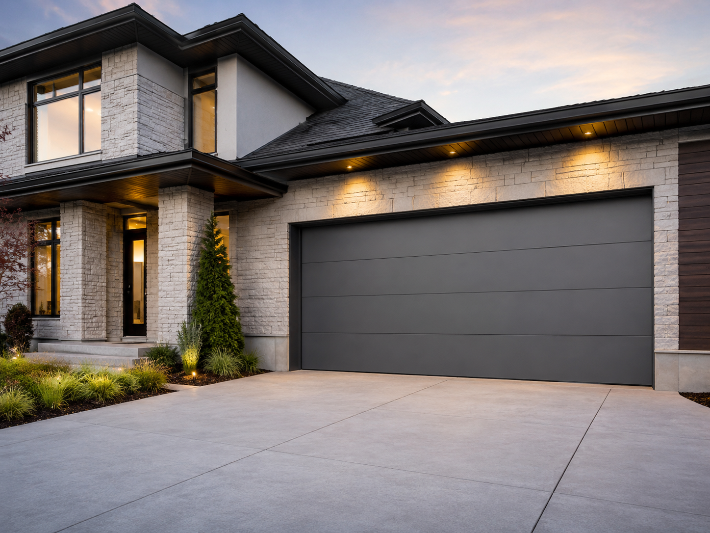 Modern two-story house with stone exterior, dark garage door, and lit exterior lights at dusk