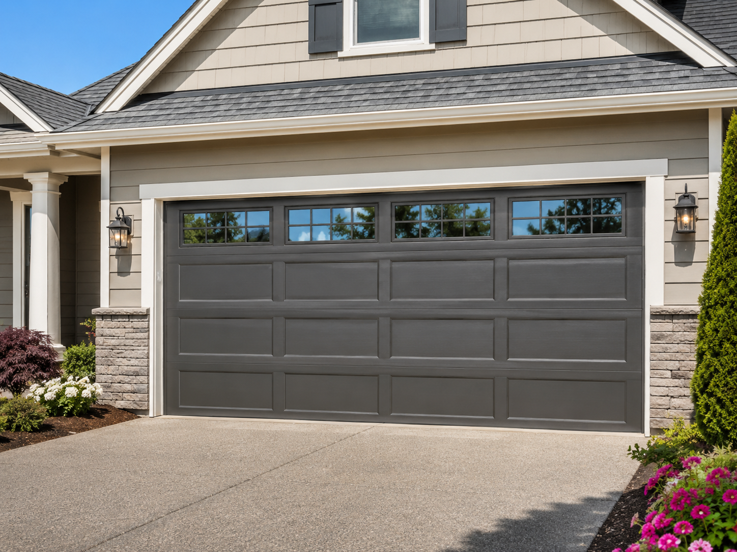 Modern suburban house front with a dark gray double garage door and driveway, framed by stone and flowers