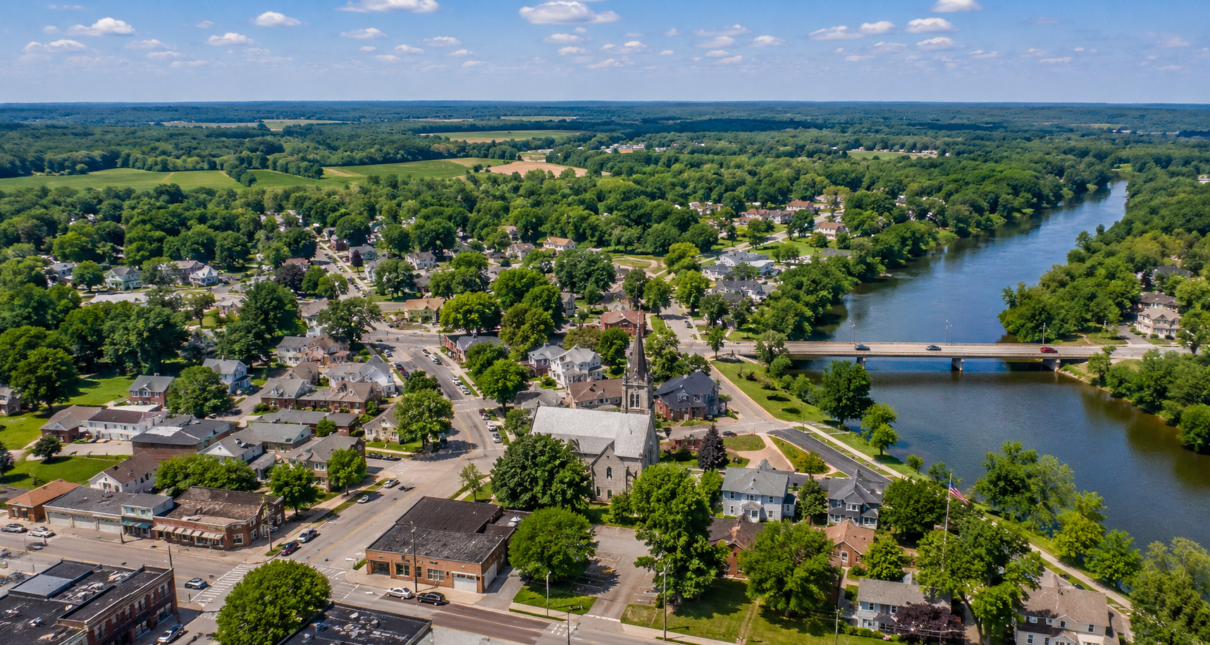 Aerial view of a small town along a river, with a bridge, houses, trees, and a church steeple.