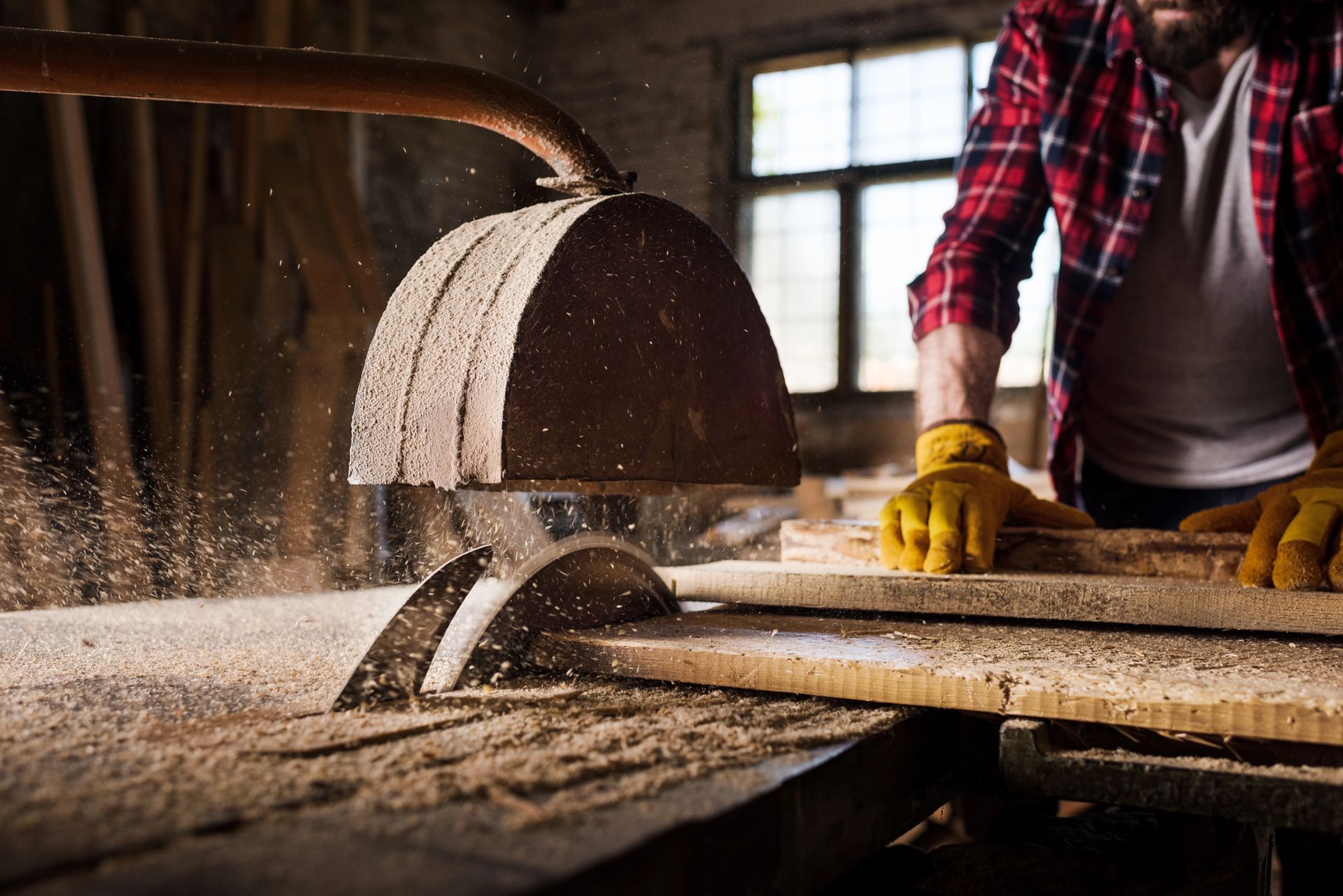 In un'officina, un falegname con una camicia a quadri rossi e guanti gialli taglia una tavola di legno con una sega circolare da banco.
