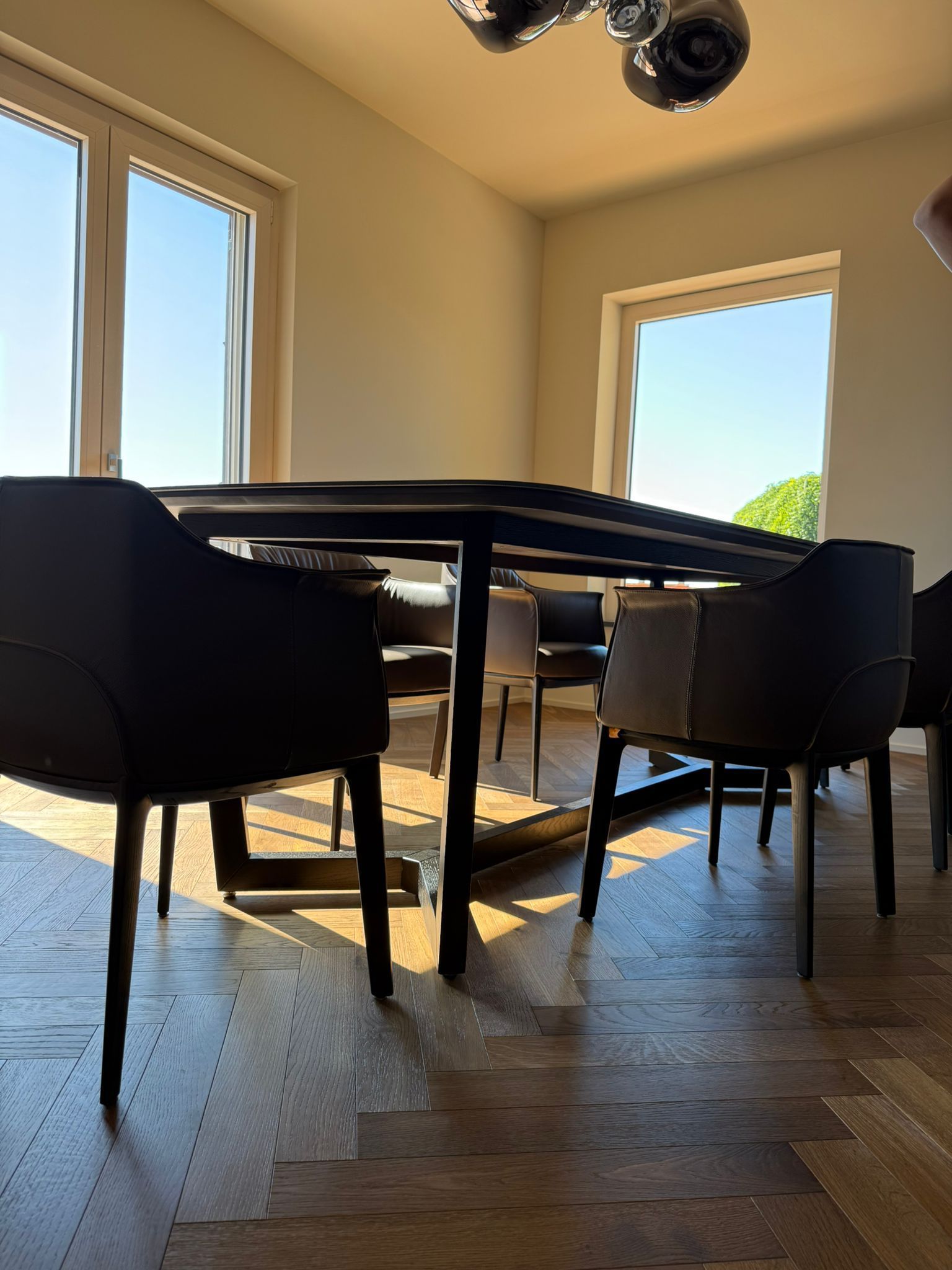 Un tavolo da pranzo moderno con sedie scure, disposto su un pavimento in legno con motivi decorativi, in una stanza luminosa con ampie finestre.