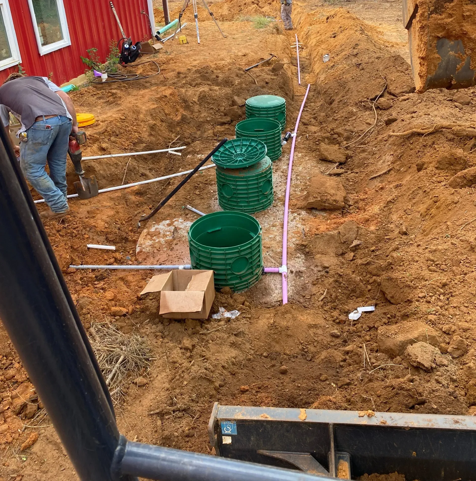 Construction trench with green utility boxes, white pipes, and a worker digging beside a red building