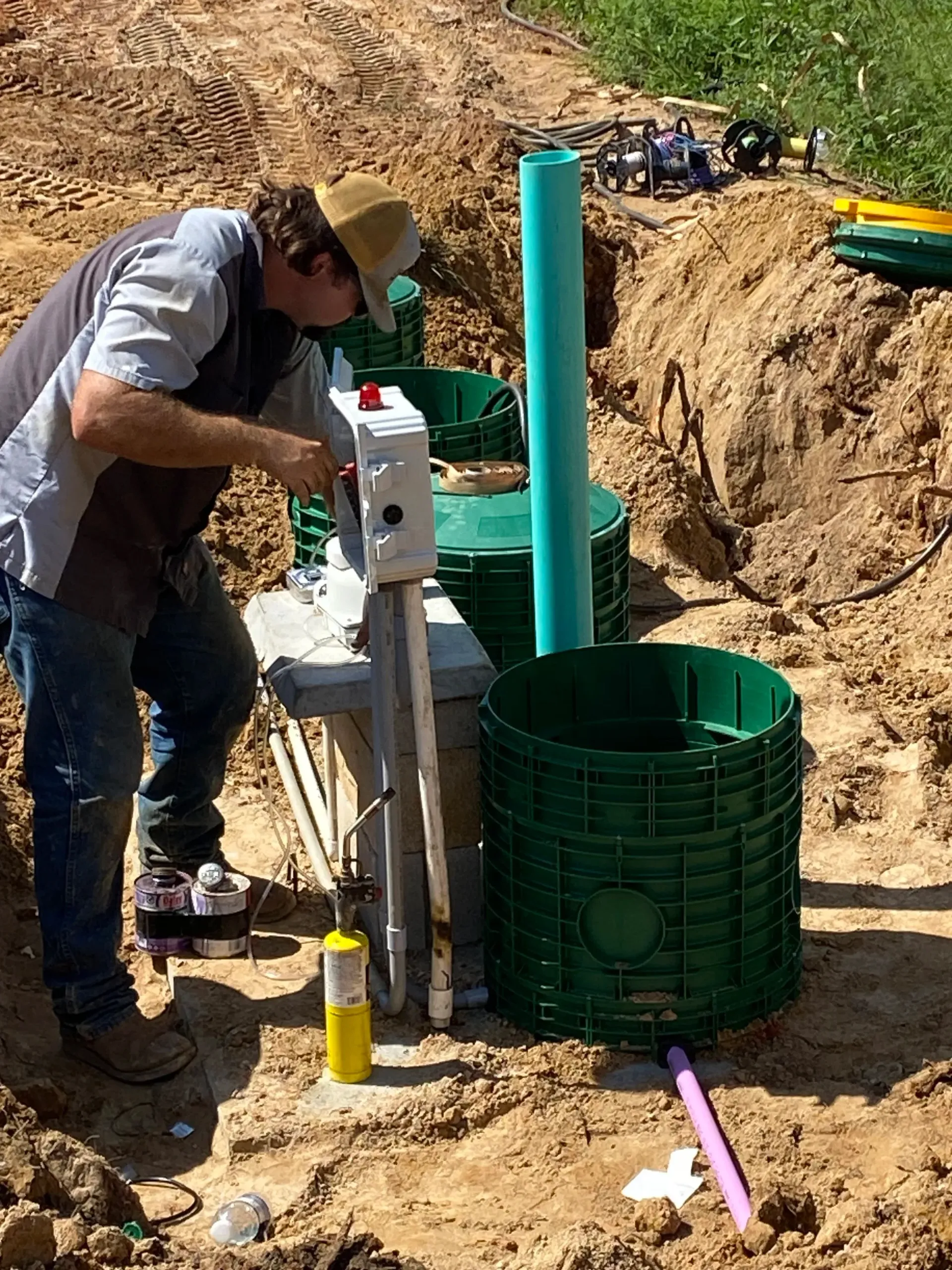 Worker using drilling equipment beside a green utility tank in a dirt trench