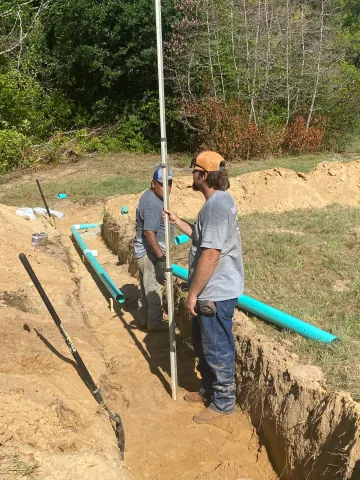 Two workers in a trench beside blue pipes, installing utility lines outdoors.