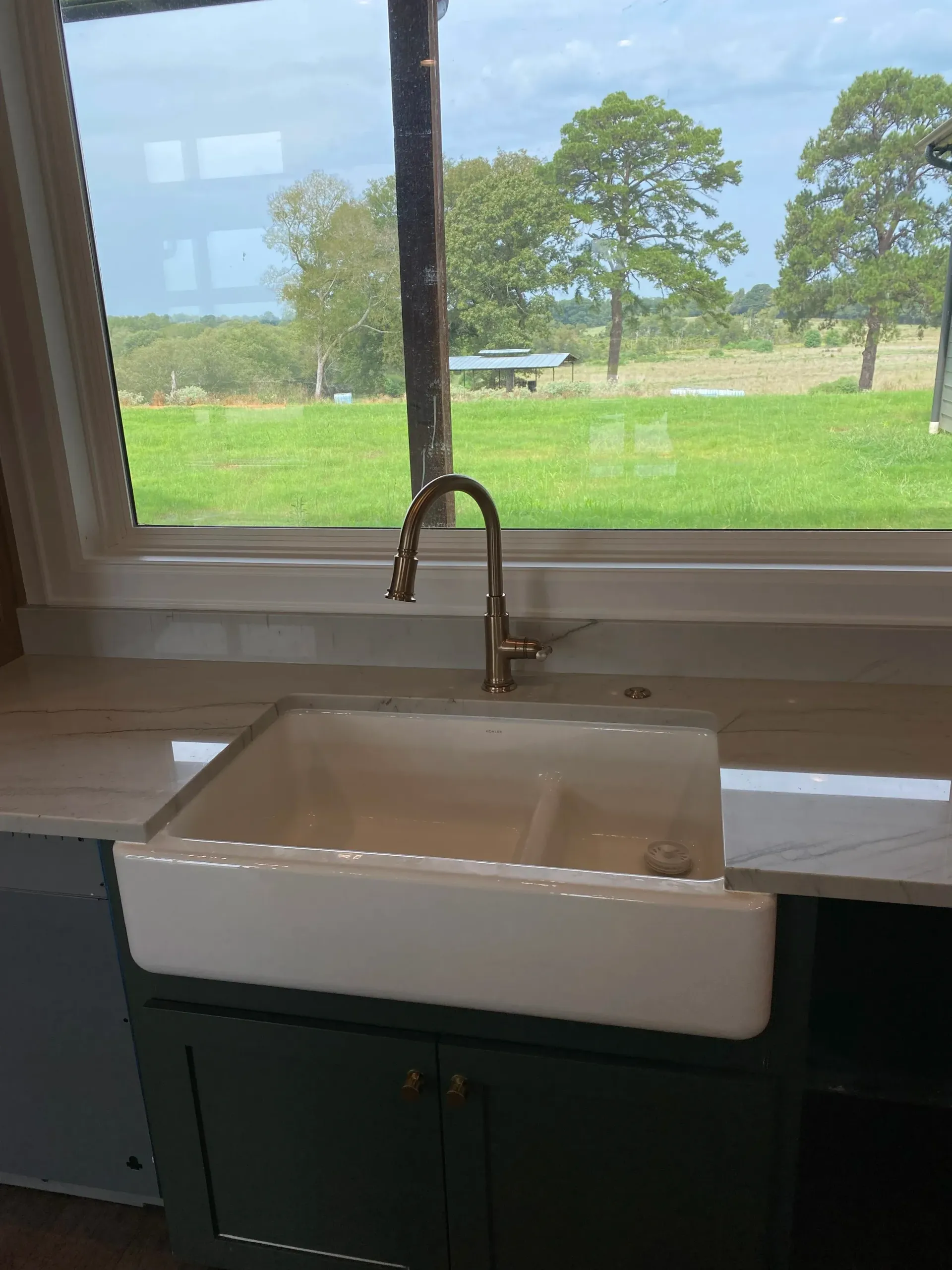 Farmhouse sink below a window overlooking a green field and trees