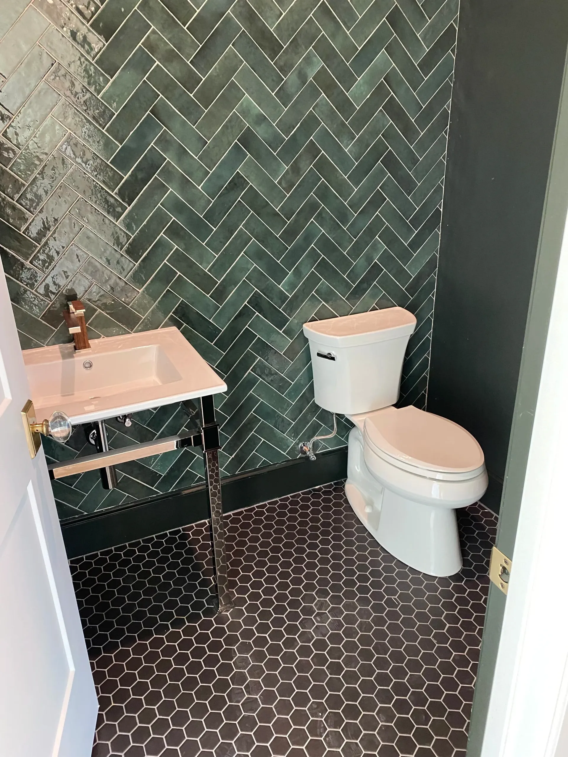 Small bathroom with herringbone green tile, white toilet, sink, and dark hexagon floor tile.