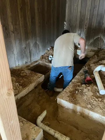 Worker in a crawlspace, digging beside concrete footings and exposed PVC pipes.