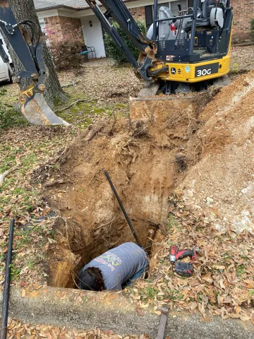 Excavator digging a trench beside a house, with a large hole and a buried pipe in the foreground.