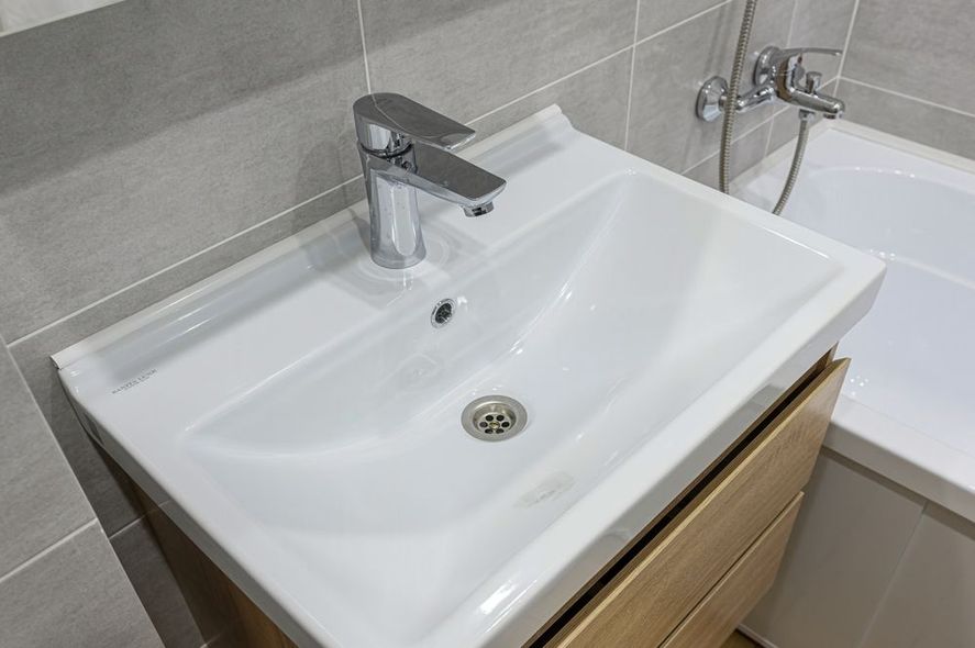 White bathroom sink with chrome faucet on a light wood vanity beside a tiled tub.