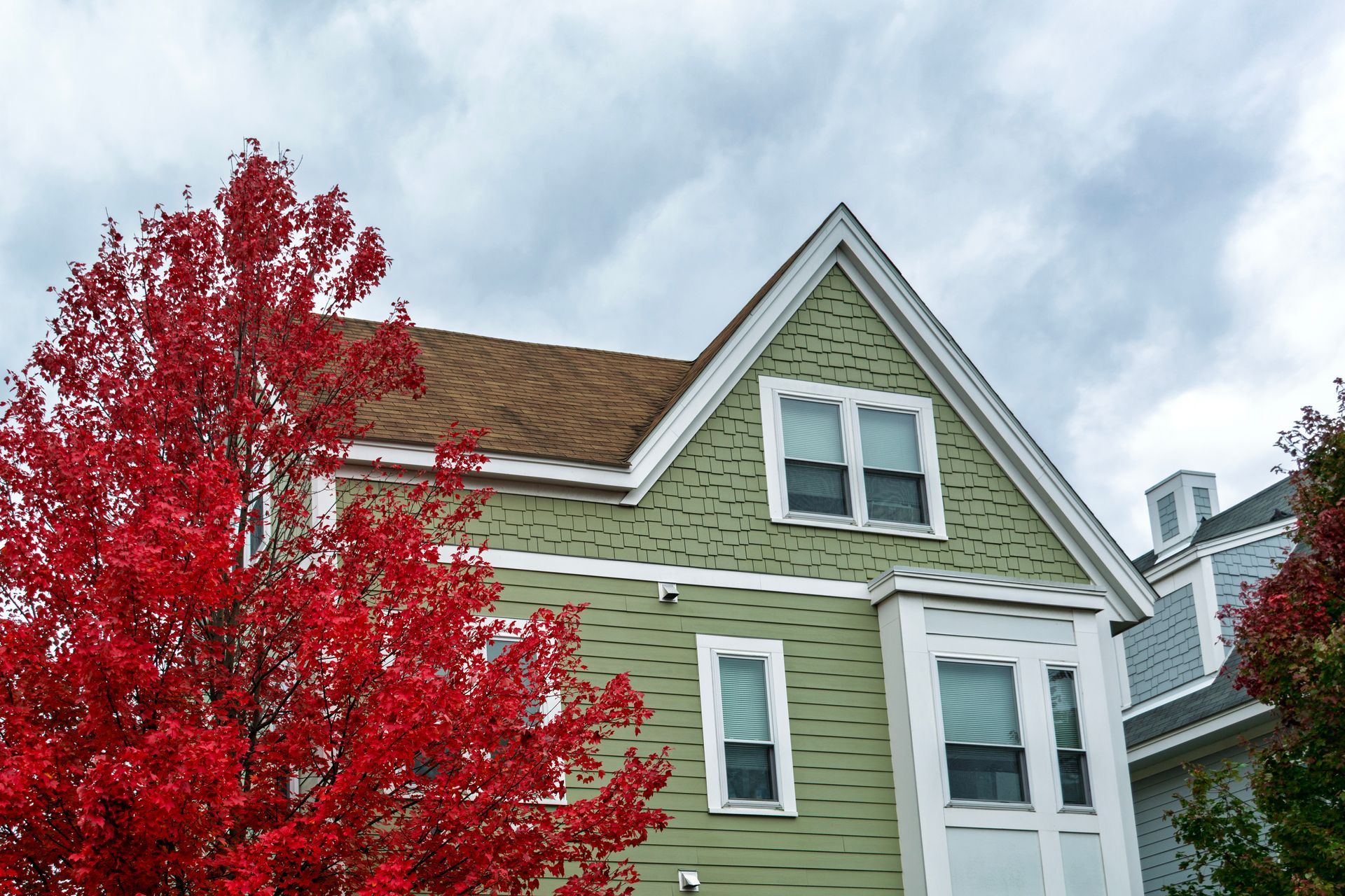 Green house with white trim and a red tree in front under a cloudy sky