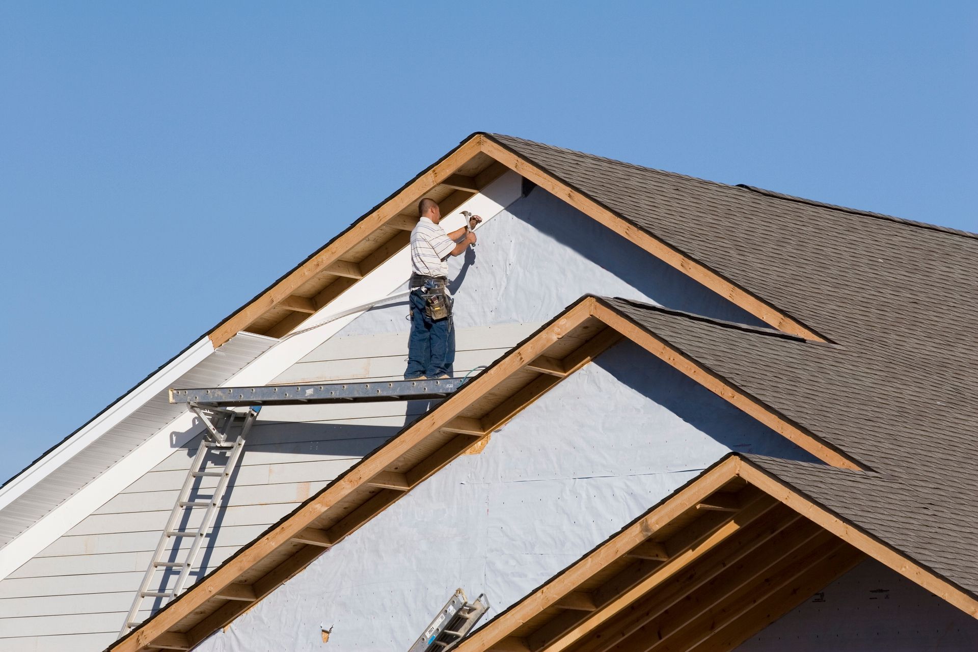 Worker repairing a roof, standing on a ladder beside gray shingles and exposed wooden framing.