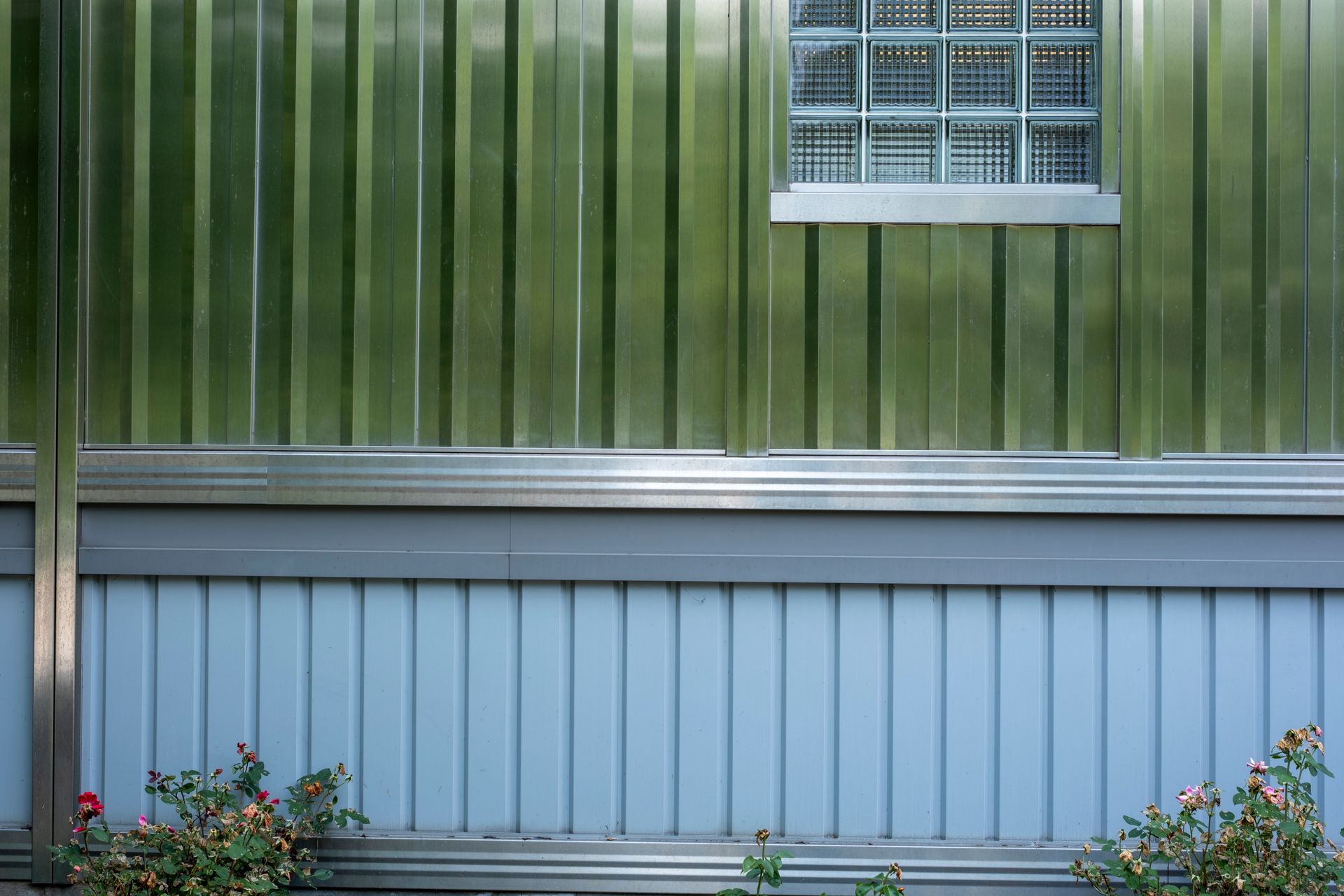 Green and blue house siding with a small window and flower beds below