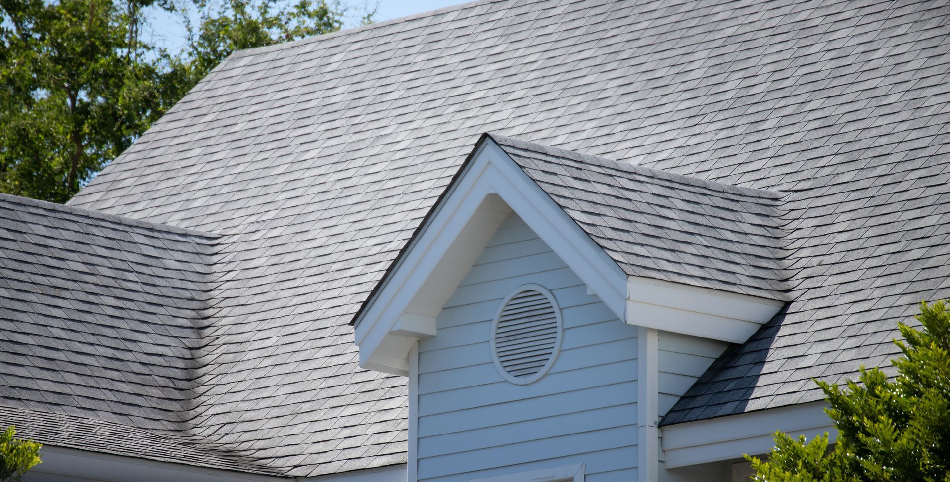 White house roof with gray shingles and a light blue dormer, framed by green trees. White house roof with gray shingles and a light blue dormer, framed by green trees.