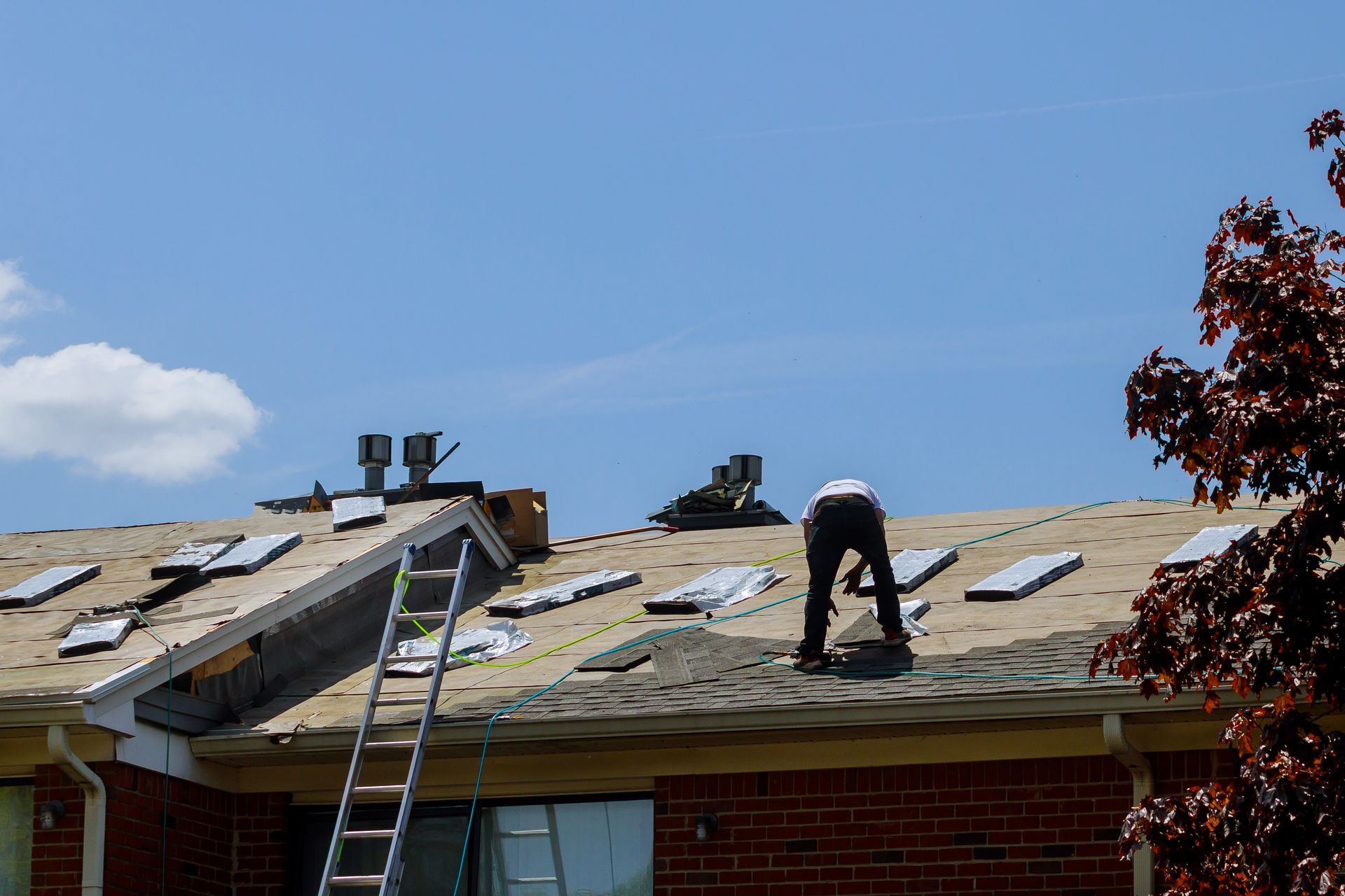 Two workers roofing a house, one on a ladder and one kneeling on the shingles under a blue sky Two workers roofing a house, one on a ladder and one kneeling on the shingles under a blue sky