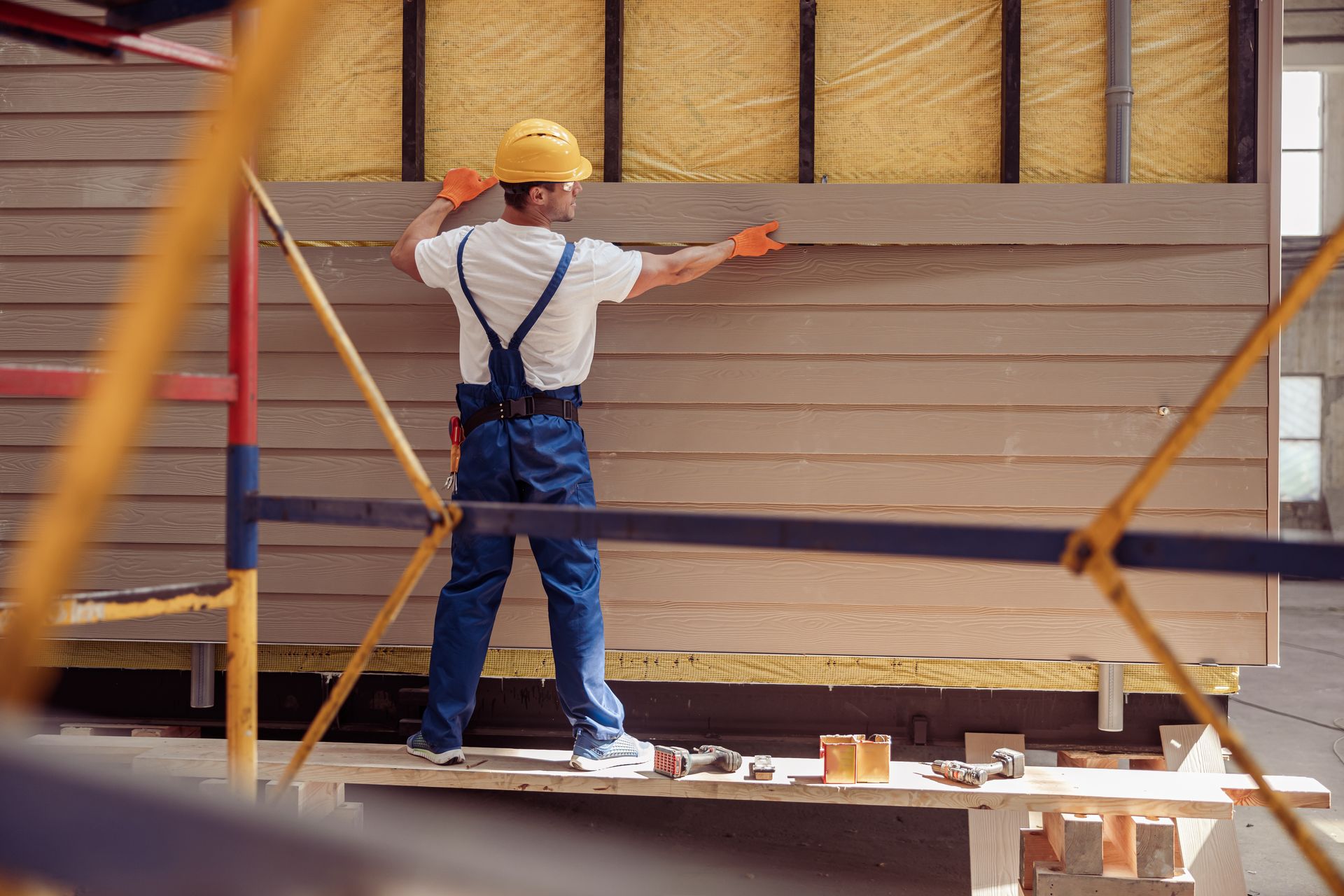 Worker in hard hat installing horizontal siding on a building frame, using tools on a scaffold.