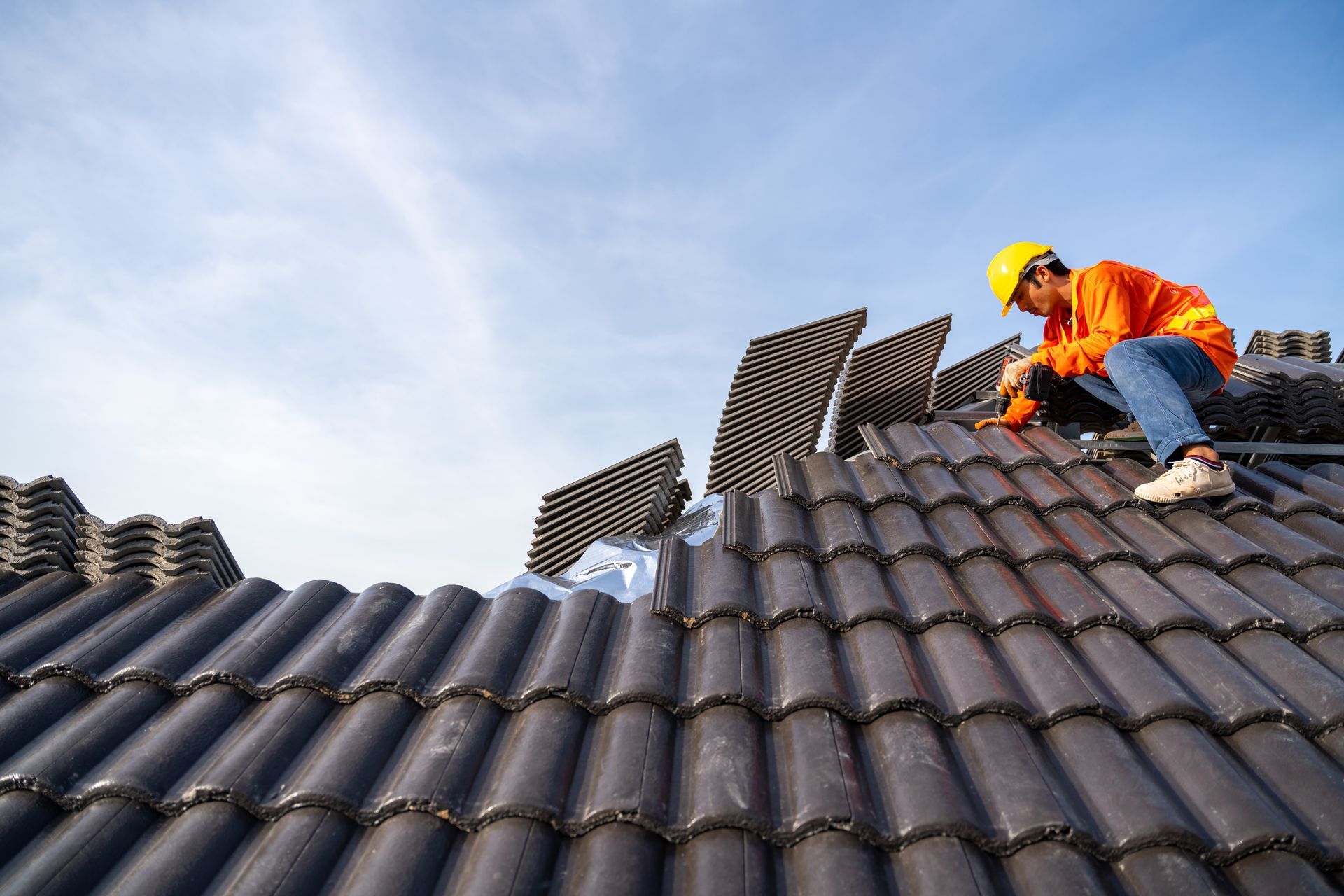 Worker in orange jacket and yellow hard hat repairing a dark tiled roof under a blue sky. Worker in orange jacket and yellow hard hat repairing a dark tiled roof under a blue sky.