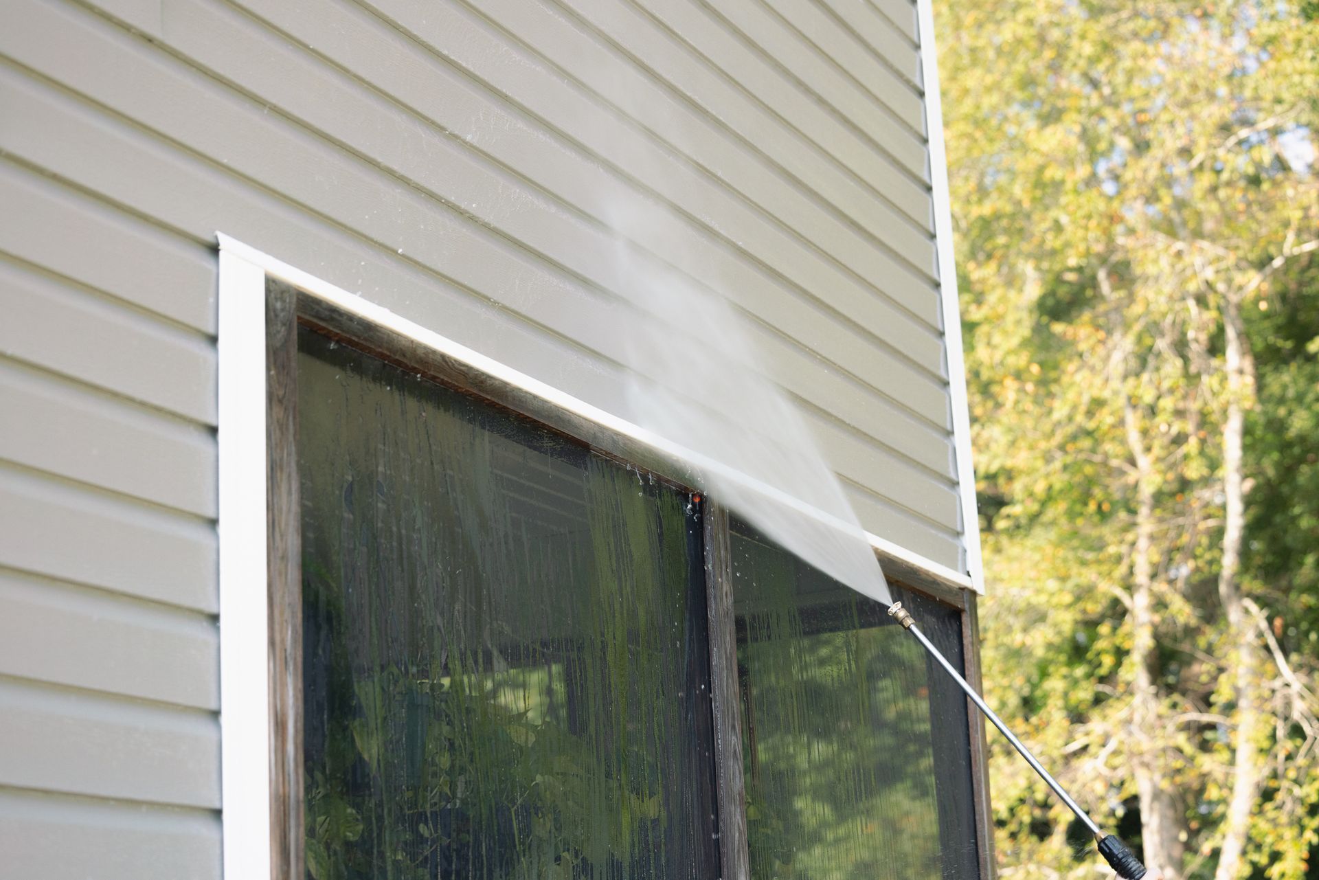 Person pressure-washing a tan house wall and window with a spray wand